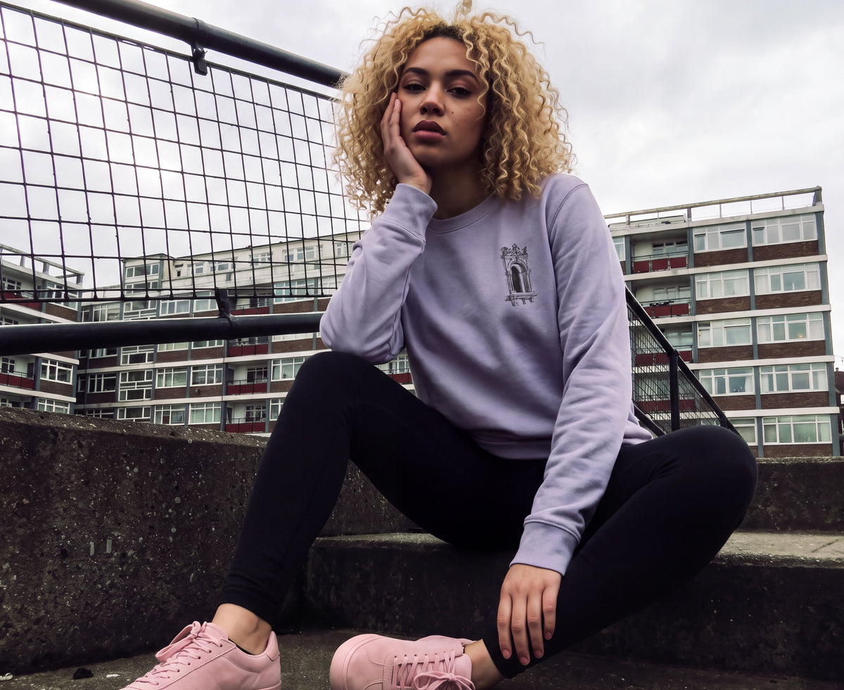 A woman with curly blonde hair sits on concrete steps outdoors, wearing a light lavender sweatshirt with a small graphic on the chest, black leggings, and pink low-top sneakers. Apartment buildings and a metal railing are visible in the background under an overcast sky.