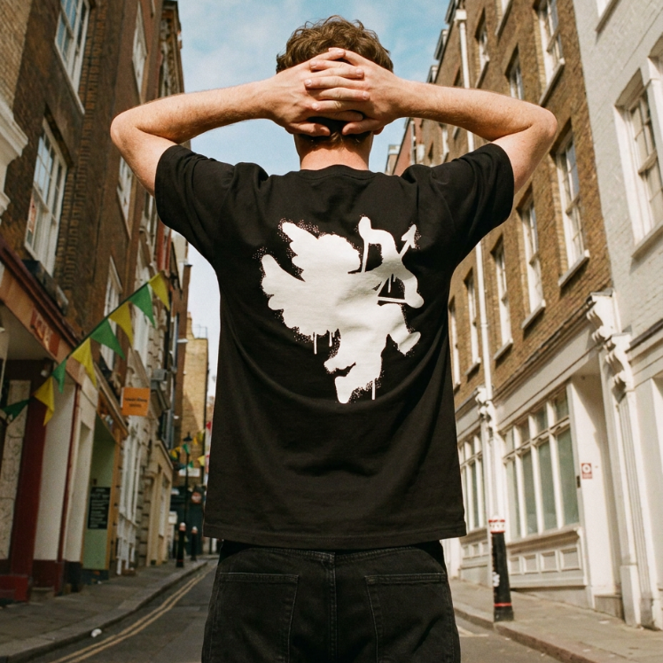 A man standing on a city street with his back to the camera, wearing a black t-shirt with a large white abstract graphic print on the back, paired with black jeans.